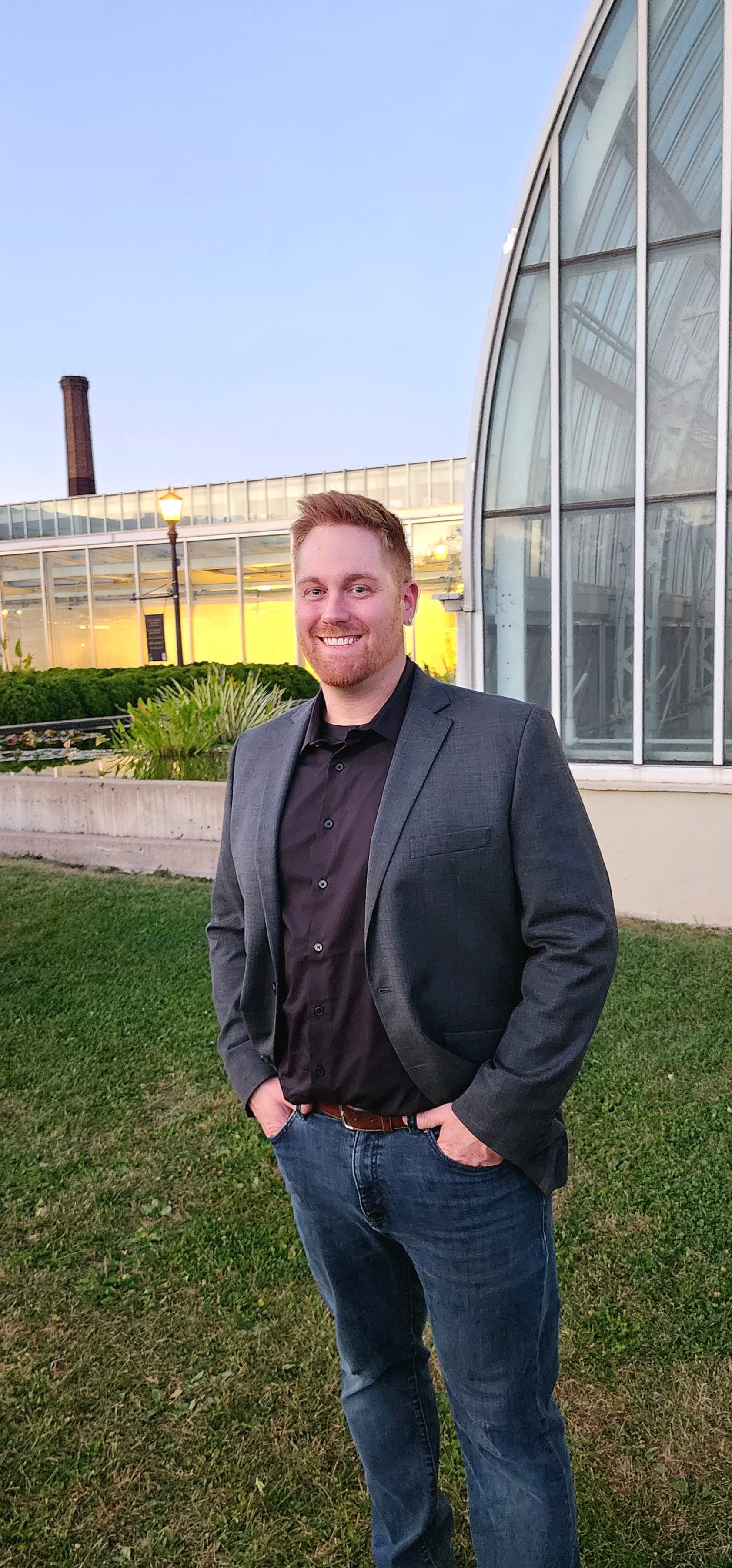 Smiling man with red hair in a grey blazer and jeans before a glass conservatory.