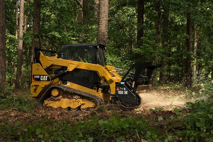 Yellow CAT compact track loader using a mulching attachment to clear brush in a forest.