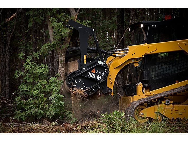 Yellow tracked skid steer with a mulching attachment grinding a tree trunk in the woods.