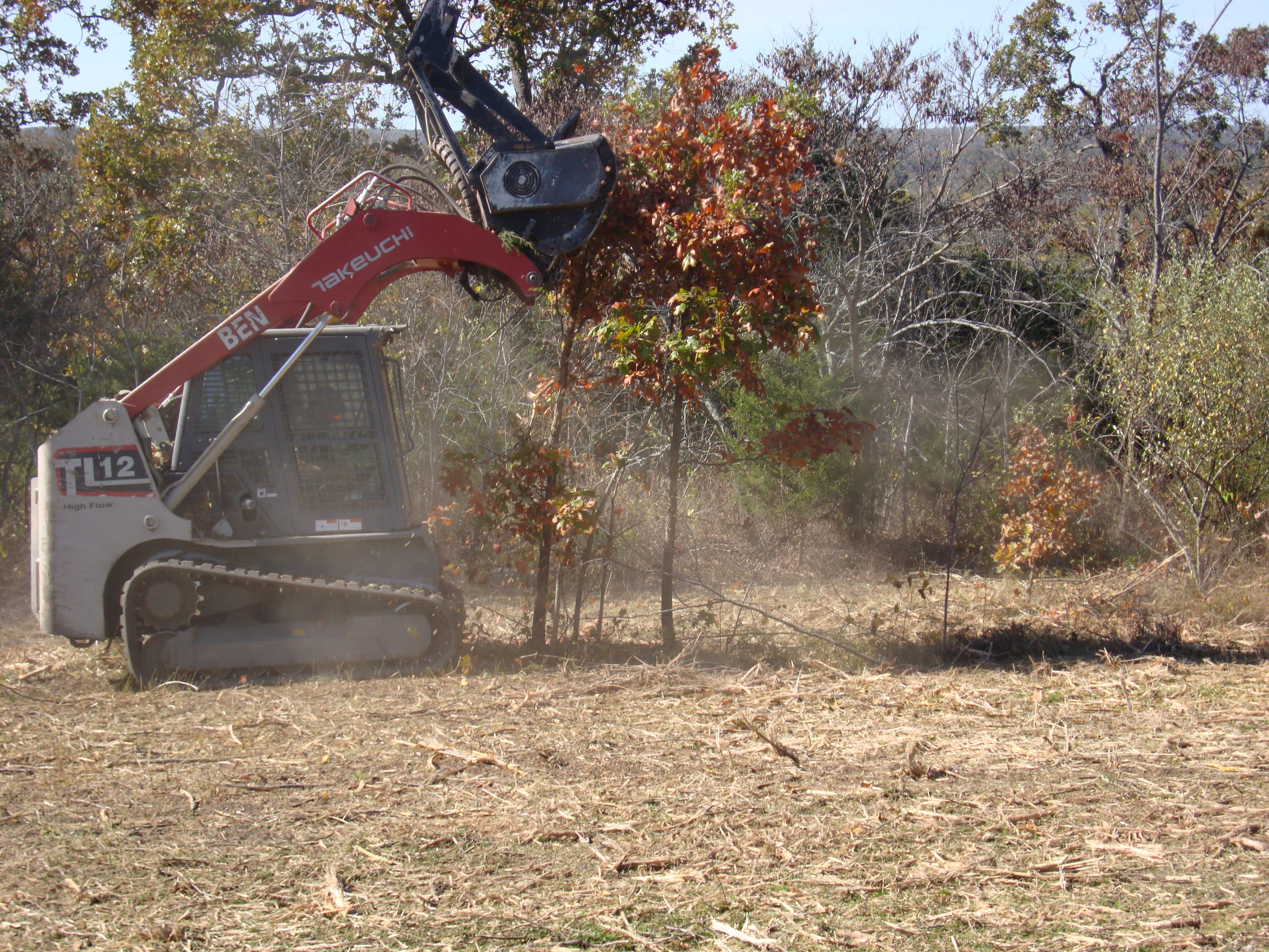 Red Takeuchi track loader mulching small trees and brush in a wooded field with flying debris.