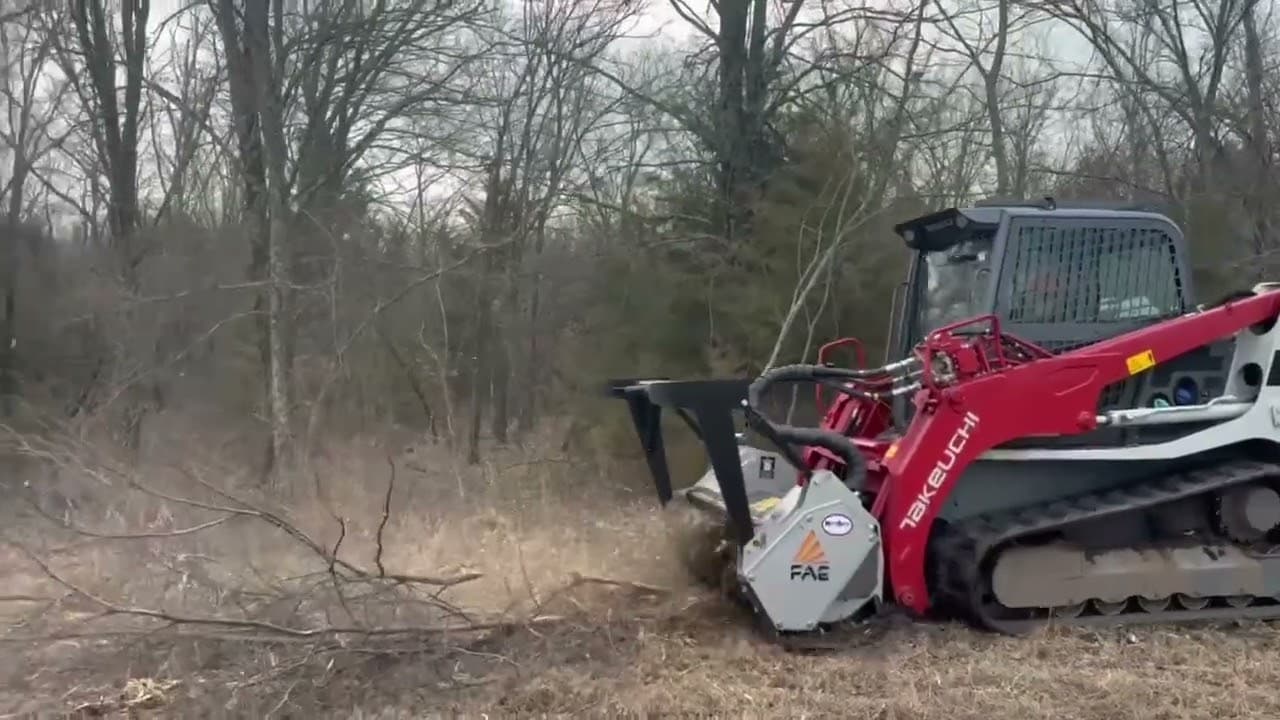 Red Takeuchi track loader with a mulcher attachment clearing brush in a wooded area.